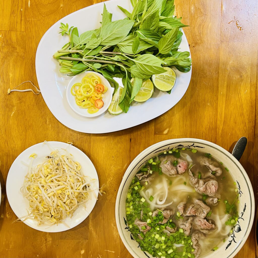 Miska z zupą Pho i talerze z dodatkami (kiełki, zioła, limonka)