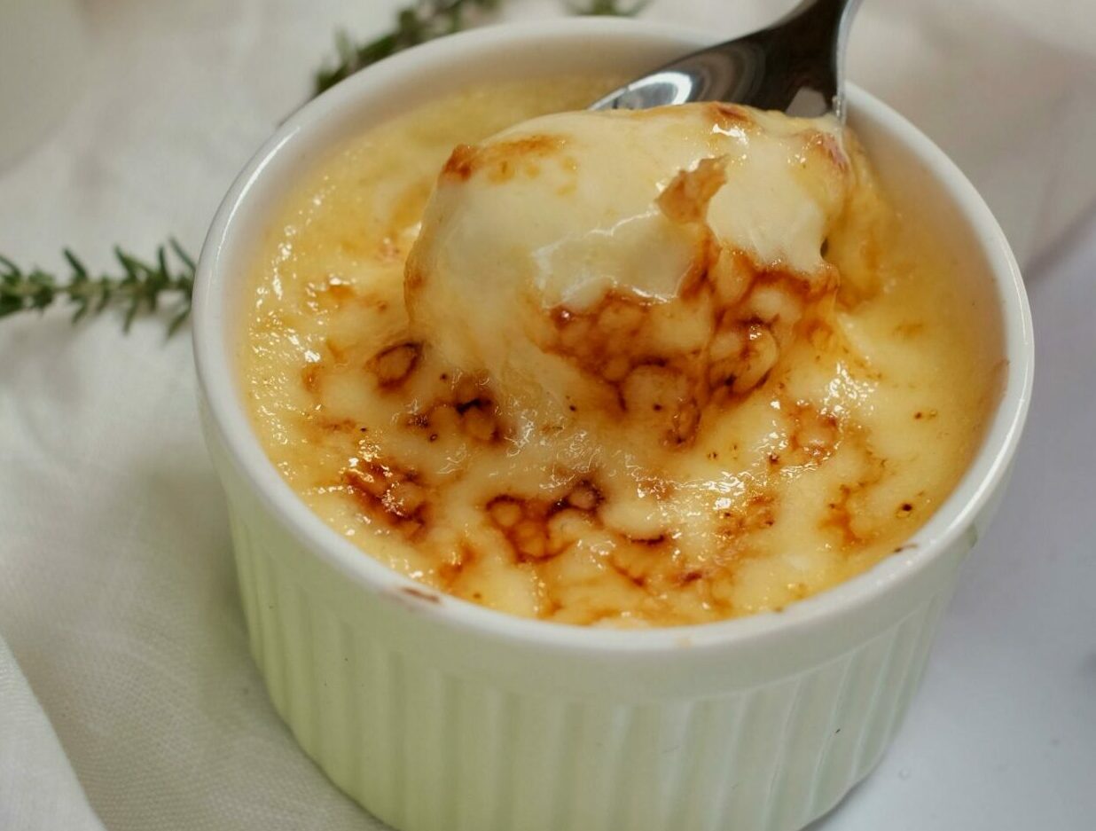 Close-up of a creamy crème brûlée dessert in white ramekin with caramelized top, garnished with herbs.
